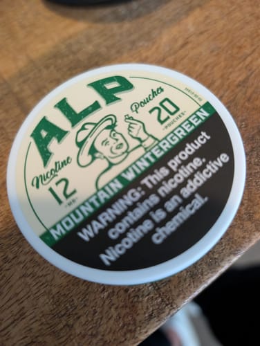 Customer's circular white can of ALP Mountain Wintergreen nicotine pouches on a wooden surface.