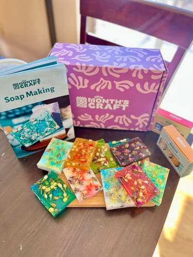 Customer shows This Month's Craft Subscription Box with the soap making guide and colorful finished soaps on a table.