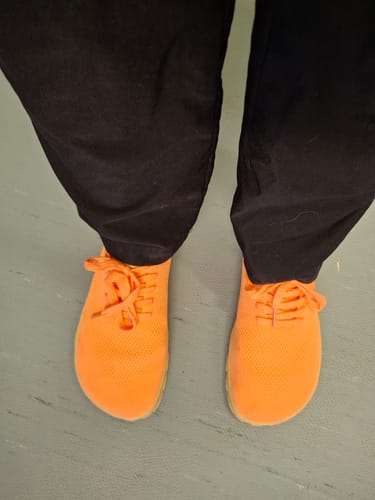 Asiakkaan kirkkaan oranssit Vaistoa Iloa -paljasjalkatennarit mustien housujen kanssa.