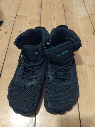 Rezensent zeigt die schwarzen, knöchelhohen Vitality V - Barfußschuhe auf einem Holzboden.