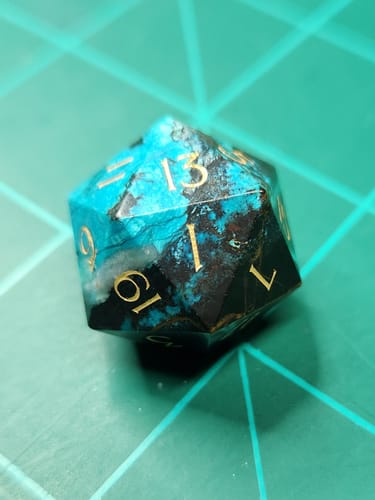 Close-up of a reviewer's turquoise and black gemstone D20 die with shiny gold numbers.
