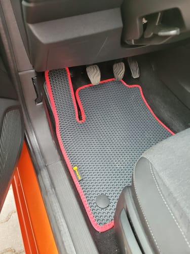 Recenzent pokazuje szary, dopasowany dywanik samochodowy z czerwoną lamówką, zainstalowany po stronie kierowcy.