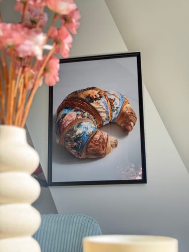 Customer's 'Un Croissant' art print in a black frame, hanging on a wall next to a vase of pink flowers.