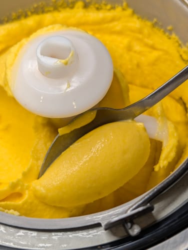 Ein Kunde schöpft cremiges, gelbes Eis aus Bio-Mangos, das in einer Eismaschine zubereitet wird.