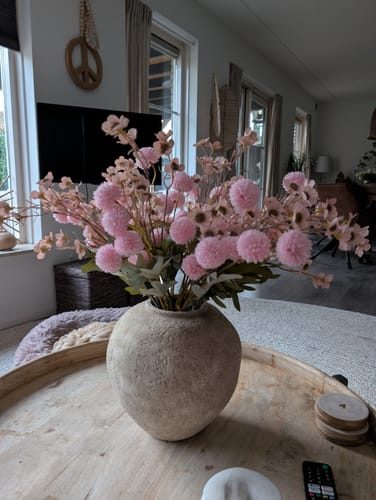 Recensent toont de beige Arda vaas op een houten tafel, gevuld met een boeket roze kunstbloemen.
