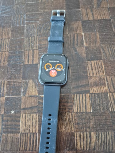 Kunde zeigt eine schwarze Smartwatch mit eingeschaltetem Display auf einer Holzoberfläche.