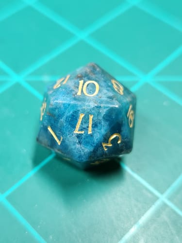 Customer's close-up of a URWizards blue apatite gemstone die with gold numbers on a green gridded surface.