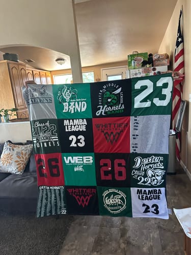 Customer's finished Project Repat T-Shirt Quilt, made from colorful school and sports-themed shirts, held up in a living room.