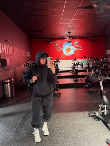 Reviewer taking a mirror selfie in a gym, wearing the oversized black Layover Hoodie with the hood up.