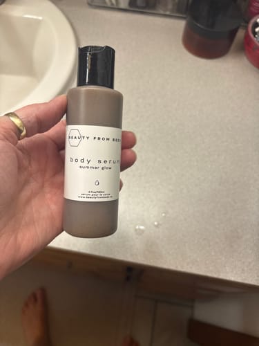 A customer holds the Body Serum - Summer Glow bottle in their hand over a bathroom counter.
