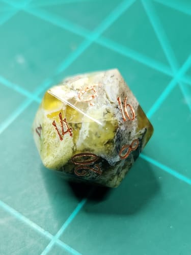 Customer's close-up of a URWizards yellow opal D20 die, showing its unique inclusions and coppery numbers on a green mat.