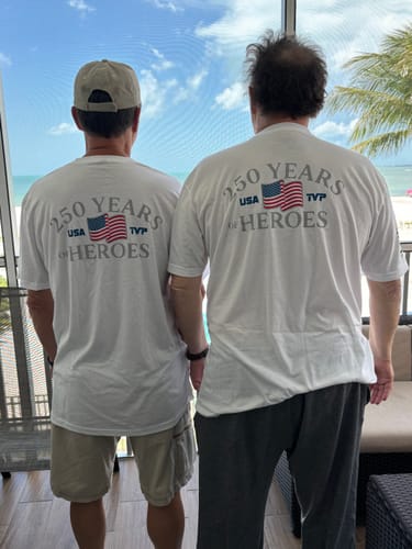 Two reviewers showing the back graphic of the '250 Years of Heroes' t-shirt while standing on a balcony.