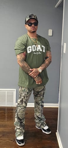Reviewer wearing a green Premium Goat One Of One Tee with a white logo, paired with camouflage pants and hat.