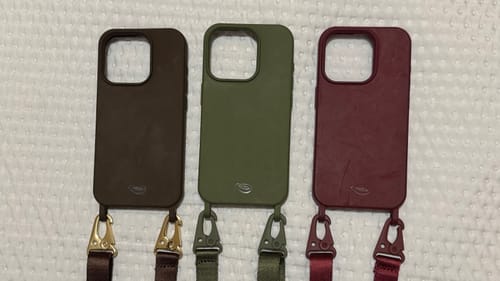 Cliente exibe três capas de celular com a Alça Aurora em bordô, verde e marrom.
