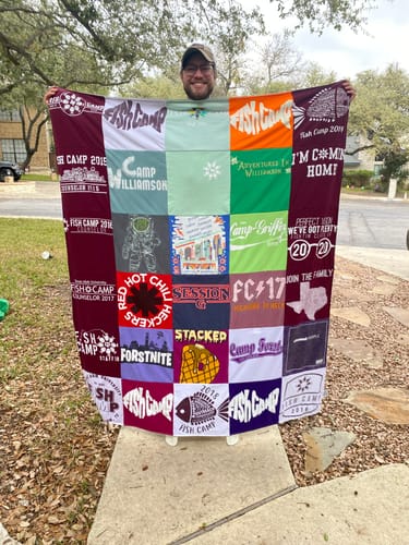 Reviewer smiling while holding up a large Premium T Shirt Quilt made of many colorful camp-themed t-shirts.