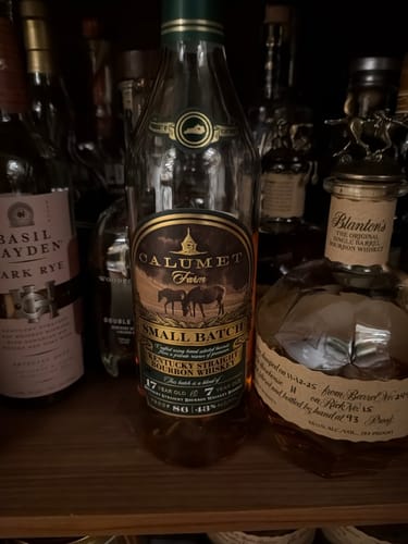 Reviewer's bottle of Calumet Farm Small Batch Bourbon displayed on a dark shelf among other whiskeys.