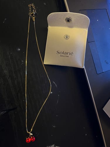 Customer's gold-colored necklace with a red cherry pendant from the VIP Crystal Club, laid out next to its white pouch.
