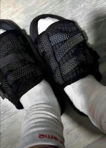 A reviewer's feet wearing black slides with patterned straps over white socks.