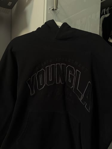 Customer's black Layover Hoodie hanging on a hanger, showing the tonal embroidered chest logo.