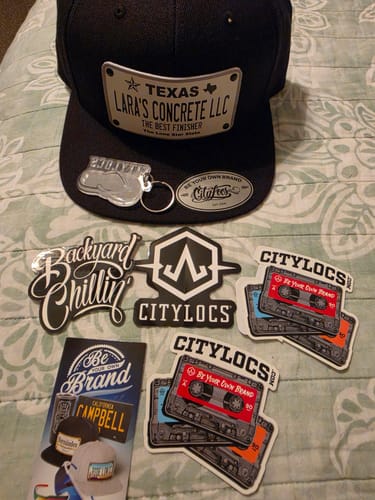 Customer's custom black Texas White Plate Hat for 'LARA'S CONCRETE LLC' displayed with branded stickers and a keychain.