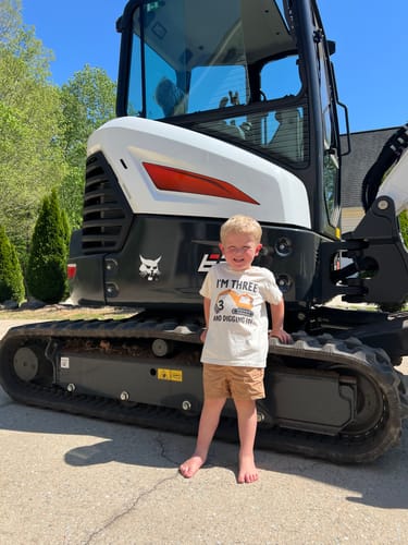 A customer's child wearing the 'I'm 3 And Digging It' shirt while smiling in front of a real excavator.