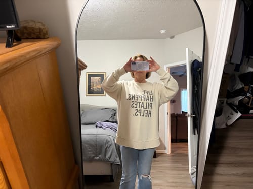 Reviewer wearing the oversized cream 'Life Happens Pilates Helps' sweatshirt in a mirror selfie.