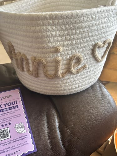 Customer's close-up of a white knitted basket personalized with the name 'Annie' and a heart icon, resting on a leather surface.