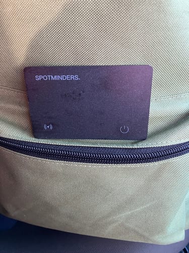 Reviewer's black SPOTMINDERS tracking card placed in the front zippered pocket of a green backpack.