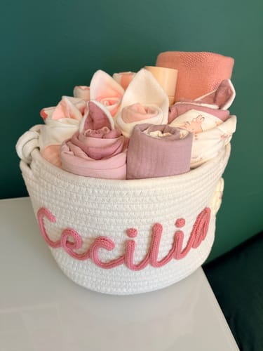Customer's white personalized knitted basket with the name 'Cecilia' in pink script, filled with rolled baby items.
