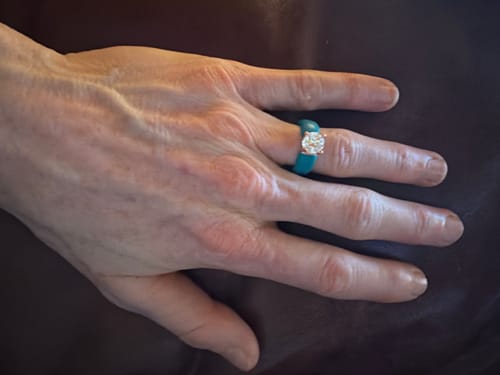 Customer's hand wearing the teal silicone ring with a round solitaire stone in a gold setting.