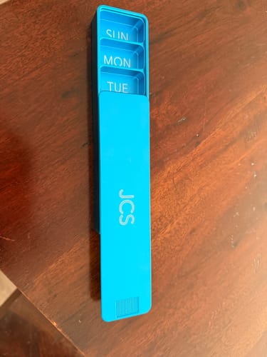 Reviewer's bright blue Weekly Case, partially open to show compartments for SUN, MON, and TUE, with 'JCS' etched on the lid.