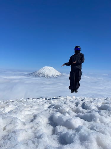 Reviewer in a black waterproof snow jacket jumping on a snowy mountain above the clouds.