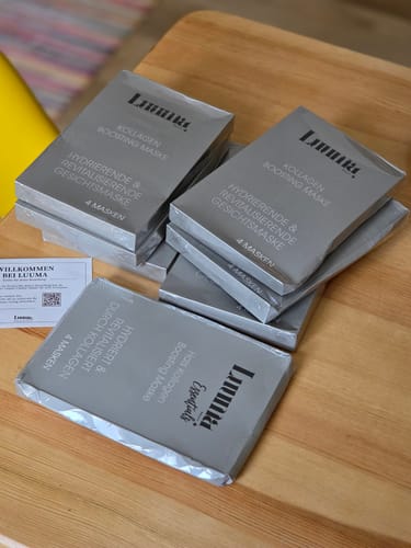 Ein Kunde zeigt mehrere graue Packungen Kollagen-Gesichtsmasken, die auf einem Holztisch liegen.