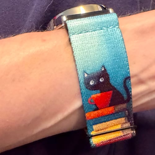 Close-up of a reviewer's Happy Here Watchband+ on their wrist, showing the black cat and books illustration.