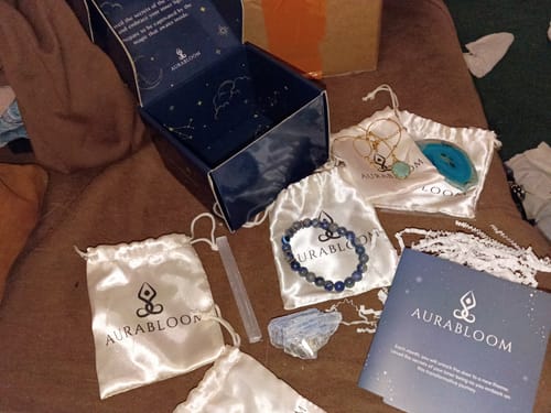 Customer displays their AuraBloom Subscription Box with included spiritual jewelry, crystals, and pouches on a couch.