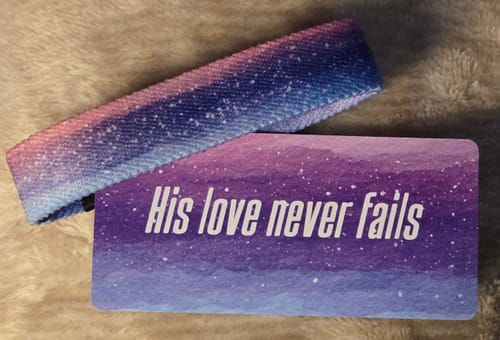 Customer's His Love Never Fails Bible Verse Bracelet with a galaxy print, shown next to its matching card on a soft surface.