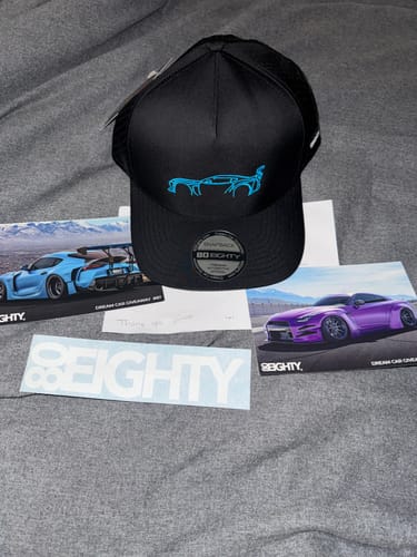 Customer's black 80Eighty DCG87 Hat with a blue car logo, displayed with branded cards and a sticker on a grey surface.