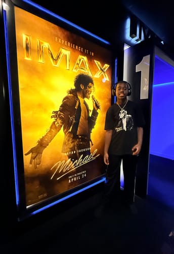 Customer wearing the black Michael T-shirt while standing next to a large movie poster for the 'Michael' movie.