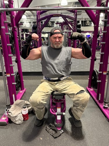 Customer flexing his biceps while sitting on a gym machine, showing the results of his strength training.