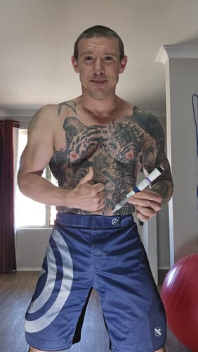 Customer with a tattooed, muscular physique holding a Genotropin 12mg pen and giving a thumbs up.