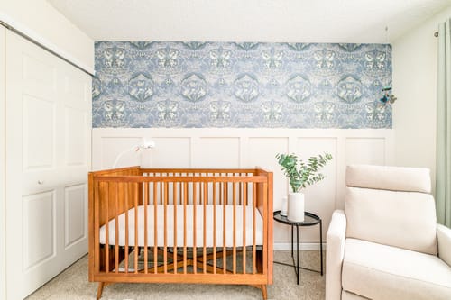 Customer's nursery with blue Cami Wallpaper on an accent wall above white wainscoting and a wooden crib.