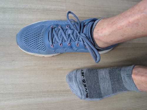 Customer's feet on a light floor, one wearing a grey striped Crew Sock and the other a blue sneaker.
