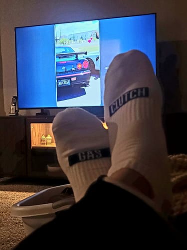 Customer relaxing with feet up, showing the 'GAS' and 'CLUTCH' print on their white ankle socks in front of a TV.