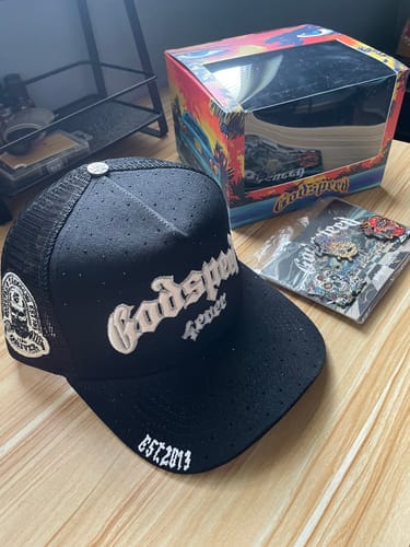 Customer's black Godspeed Forever VVS Trucker Hat displayed on a wooden table next to its original box and some pins.