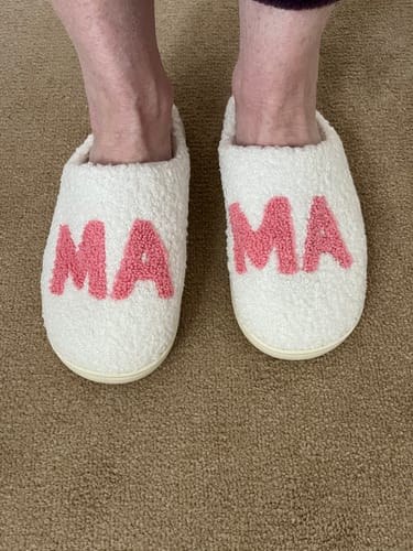 Customer wearing the plush white Mom Essential Slippers with pink "MAMA" text on a brown carpet.