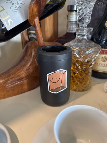 Customer's smiley paper bag sticker from the sticker pack applied to a black can cooler on a counter.