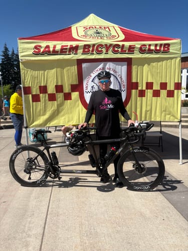 Reviewer wearing the black SOLO MIO Logo T-Shirt while standing with a tandem bicycle at an outdoor event.