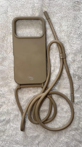Cliente mostrando a Cordinha Odara na cor areia. A capa de celular e a alça removível estão em uma superfície felpuda.