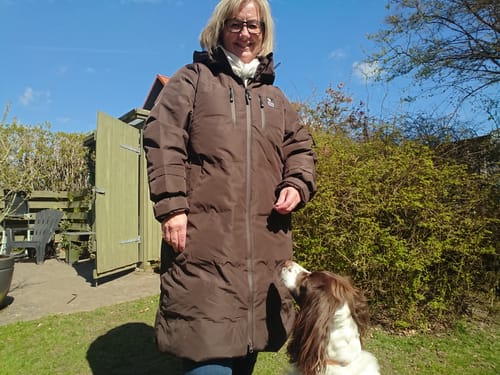 Anmelder i fuld figur iført den lange, brune KeepDry Frakke i en have med en hund.