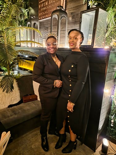 A reviewer wearing the black long-sleeved asymmetric maxi dress with gold buttons, standing indoors.